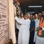 Bihar's Chief Minister Nitish Kumar inaugurating the 'Bapu Tower' in Patna, placing flowers at Mahatma Gandhi’s statue with the towering structure in the background. The modern tower, surrounded by greenery, stands tall with dignitaries and spectators celebrating the event. A tree-planting ceremony also takes place near the tower, emphasizing environmental sustainability.